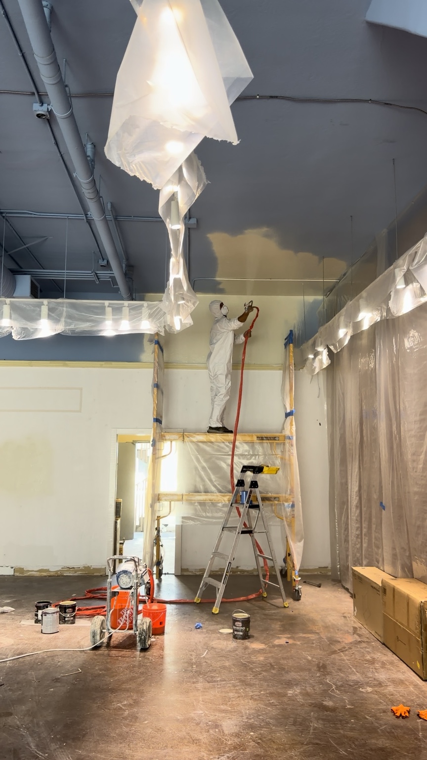 A worker spray-painting a tall ceiling in a prepared commercial room